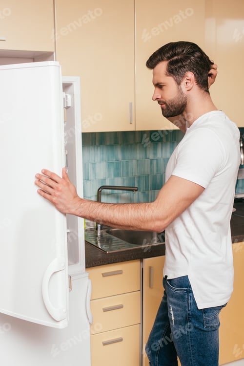 Preview: Side view of pensive man looking at open fridge in kitchen