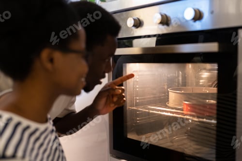 Preview: Black couple baking cake for special occasion together at home, looking inside oven with impatience
