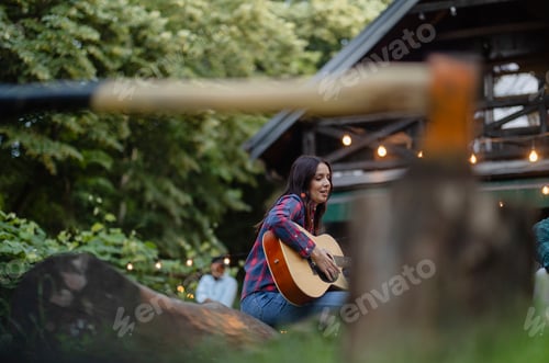 Preview: Woman playing guitar and singing at a forest party near wooden house