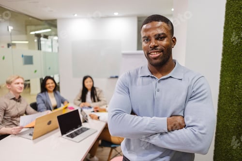 Preview: Confident businessman smiling with folded arms in modern office with coworkers meeting