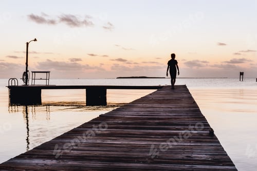 Preview: Silhouette of a teen boy walking along a dock at dusk by the ocean against a cloudy sky.