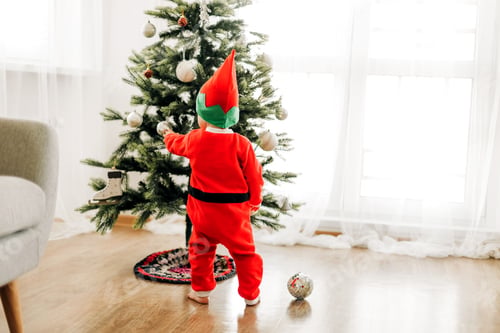 Preview: A little boy kid in a red suit stands near a decorated Christmas tree. Rear view.