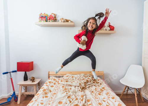 Preview: Cute happy girl in a red sweater jumping on the bed during winter holidays