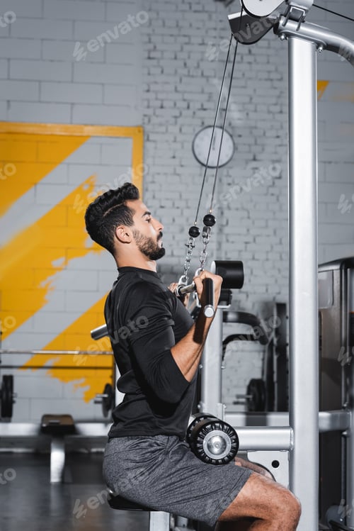 Preview: Side view of muslim sportsman training with lat pulldown machine in sports center