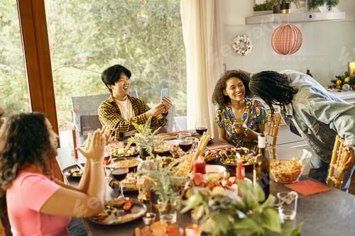 Preview: Cheerful man making photo on phone for smiling friends during festive home party