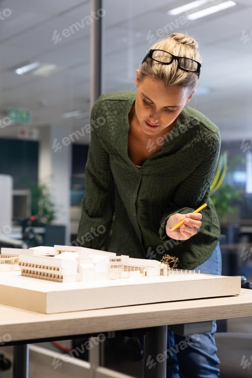 Preview: Caucasian female architect analyzing model on desk with pencil at modern workplace