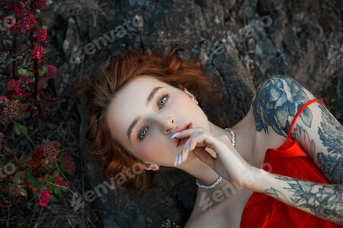 Preview: young woman with bold tattoos lies on rocks, gazing thoughtfully at the camera