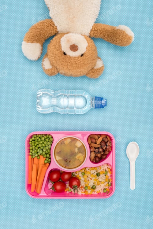Preview: top view of tray with kids lunch for school, teddy bear and bottle of water on blue surface