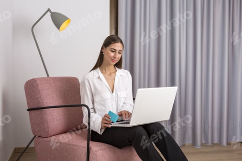 Preview: Smiling woman making purchase online on laptop sitting in armchair
