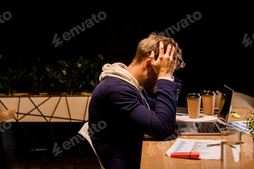 Preview: young exhausted businessman hollding hands on head while sitting at night in office near laptop and
