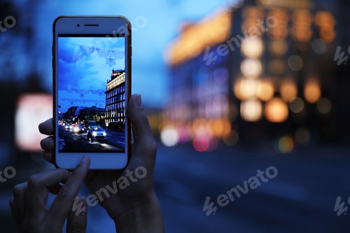 Preview: Woman taking photo of modern city in evening with smartphone, focus on hand. Bokeh effect