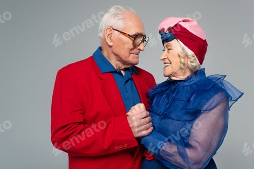 Preview: smiling elderly man in red blazer holding hands with wife in blue dress and turban isolated on grey