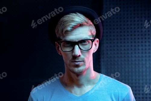 Preview: Serious young man in blue pullover, black hat and eyeglasses standing in studio