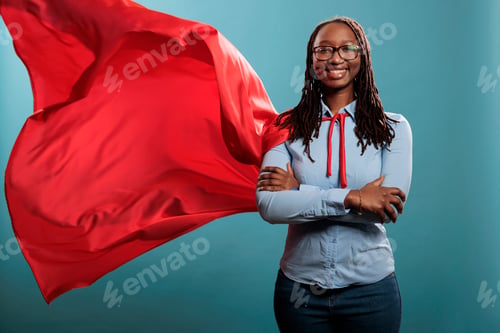 Preview: Brave and happy looking young adult superhero woman wearing red hero cape while smiling at camera.