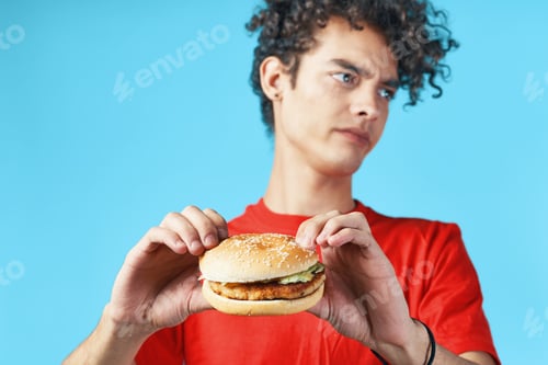 Preview: Cheerful guy with curly hair in a red t-shirt with a hamburger in his hands fast food diet