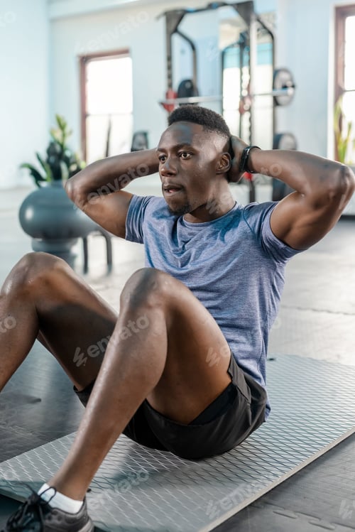 Preview: African American adult male performing crunches on gray mat in gym by barbell, wearing blue shirt