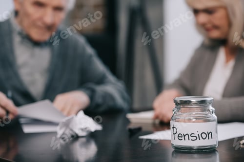 Preview: selective focus of empty glass jar with 'pension' lettering on table with senior couple on