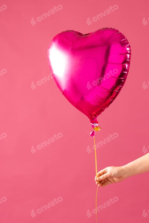 Preview: Vertical of hand holding shiny pink heart shaped balloon on pink background with copy space