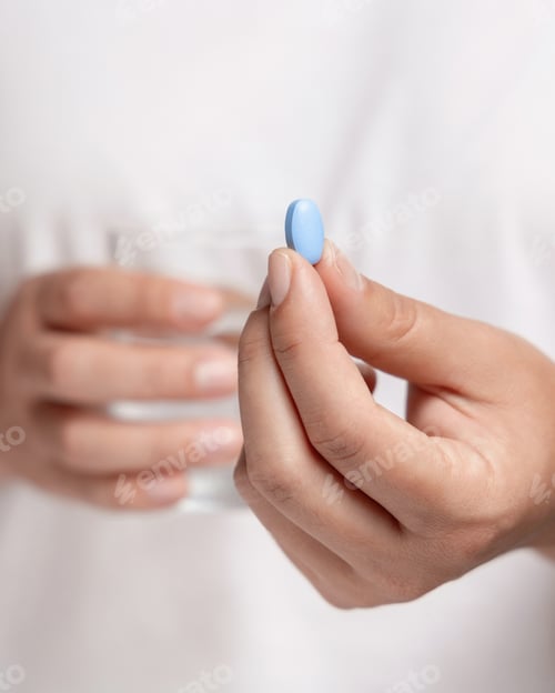 Preview: Woman in white tee holding blue pill in fingers and water glass in hand closeup. Healthcare concept