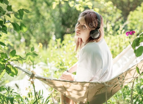 Preview: Teenager relaxing in hammock listening to music with headphones in lush green garden