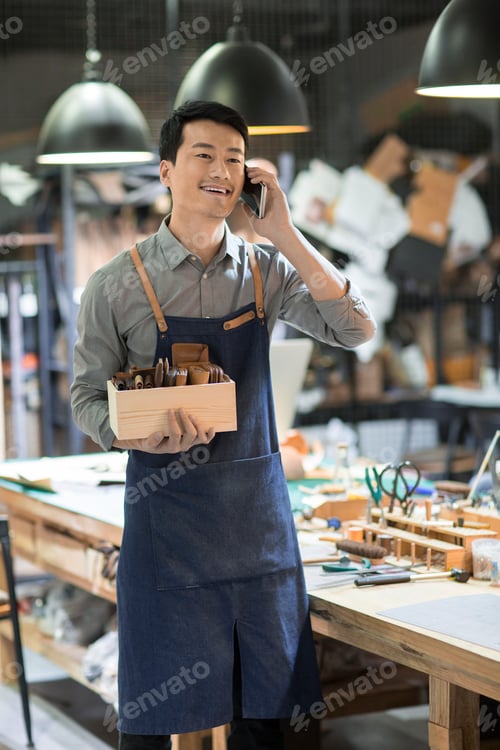 Preview: Young Chinese leather craftsman talking on cell phone in studio