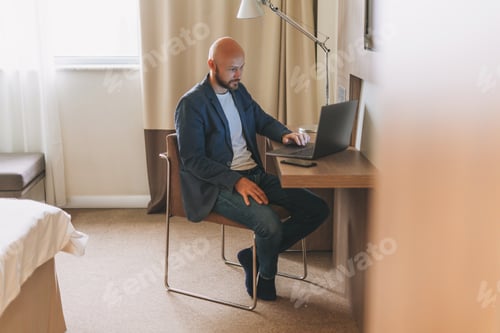 Preview: Adult bald bearded man in blue jacket with coffee working on laptop in hotel room