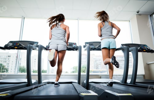 Preview: Two young beautiful slim women in sportswear running on treadmills in gym with windows and city at