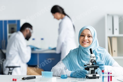 Preview: Positive muslim scientist looking at camera near microscope, flask and pipette in lab