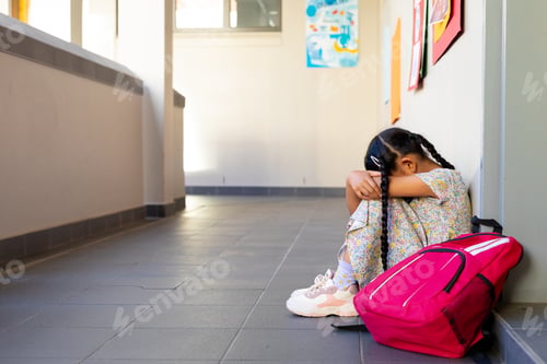Preview: Sad biracial schoolgirl with school bag sitting at wall at elementary school corridor, copy space