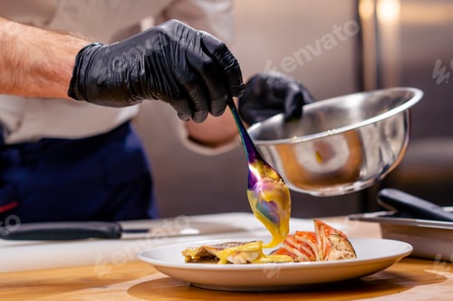 Preview: close up chef in a white tunic holding a bowl and pouring sauce from spoon onto the finished dish