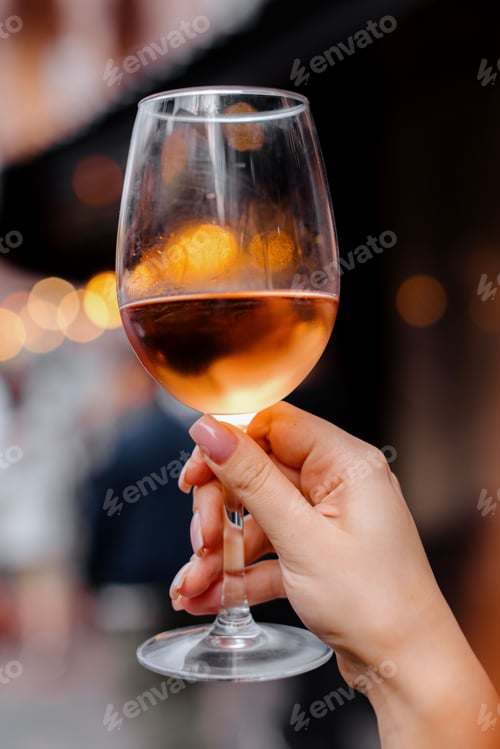 Preview: Hand holding a glass of rose wine with blurred lights.