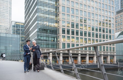 Preview: Businessman and businesswoman using mobile phone and pulling trolley luggage, Canary Wharf, London,