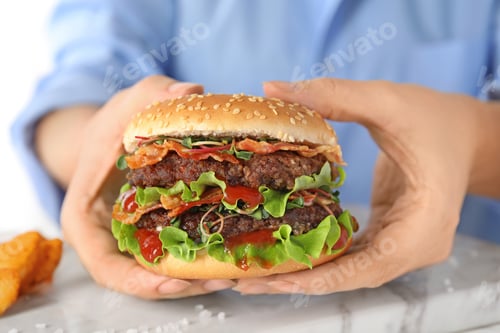 Preview: Woman holding tasty burger with bacon at table, closeup