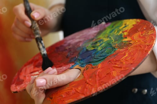Preview: Woman with paintbrush mixing paints on palette against blurred background, closeup
