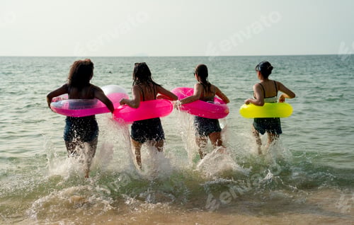 Preview: Back of teen girls with swimming rings run in the sea with warm light of sunset during holiday