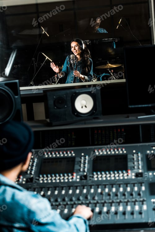 Preview: selective focus of mixed race sound producer working at mixing console while pretty woman singing in