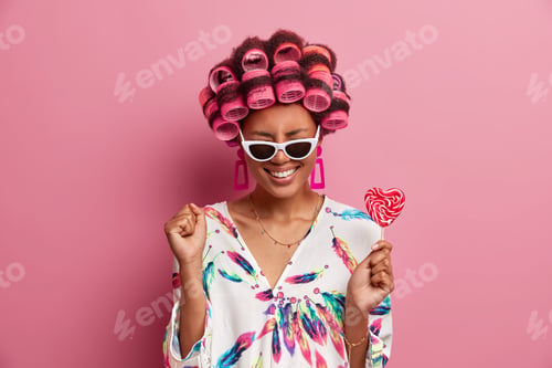 Preview: Overjoyed ethnic woman smiles broadly and clenches fist prepares for special occasion wears hair rol