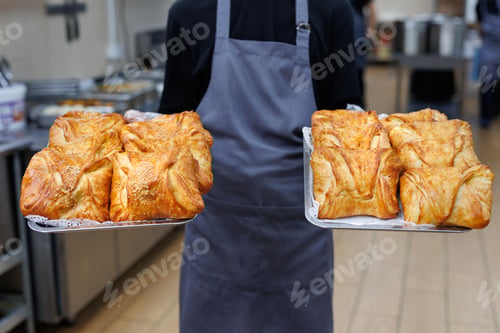Preview: Baker in apron holding two trays of freshly baked khachapuri made from puff pastry with cheese.