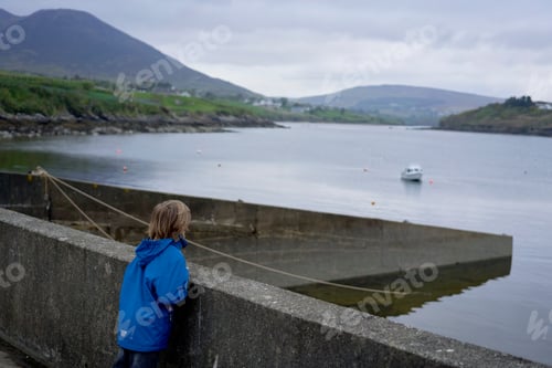Preview: Child in blue jacket looking out at a calm bay with a boat and distant hills under an overcast sky.