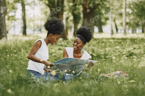 Preview: Young african-american twin sisters sitting on a grass in a park and looking in a map