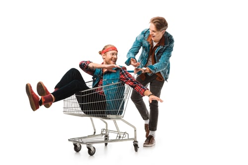 Preview: smiling young man carrying shopping cart with male friend gesturing by hands isolated on white