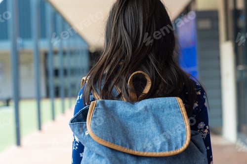 Preview: Woman with long dark hair carries a blue denim backpack, standing in a school corridor