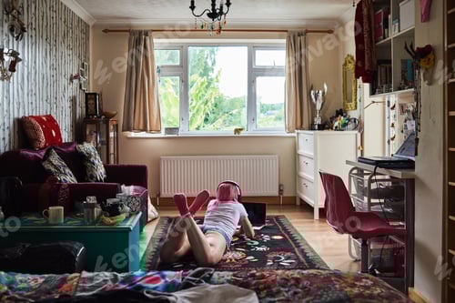 Preview: Young woman with pink hair lying on carpet wearing headphones and using laptop at home