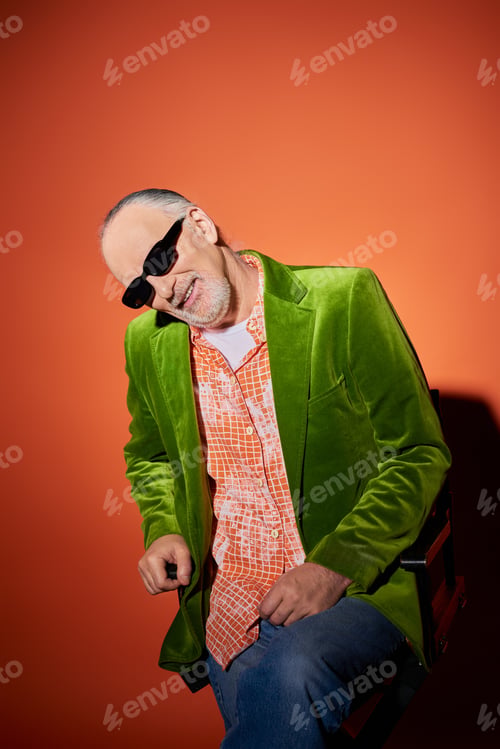 Preview: joyful senior man with grey hair and beard sitting on chair and smiling at camera on red and orange