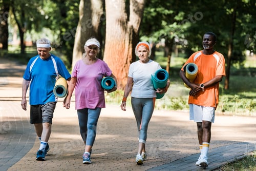 Preview: happy senior and multicultural pensioners holding fitness mats and walking in walkway in park