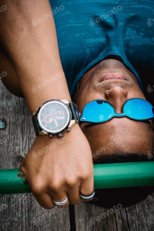 Preview: Young fit man with sunglasses and a blue shirt working out in the park while listening to music