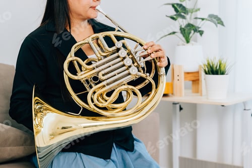 Preview: A woman sits on a cozy sofa in her living room, playing a shiny gold French horn. The warm lighting