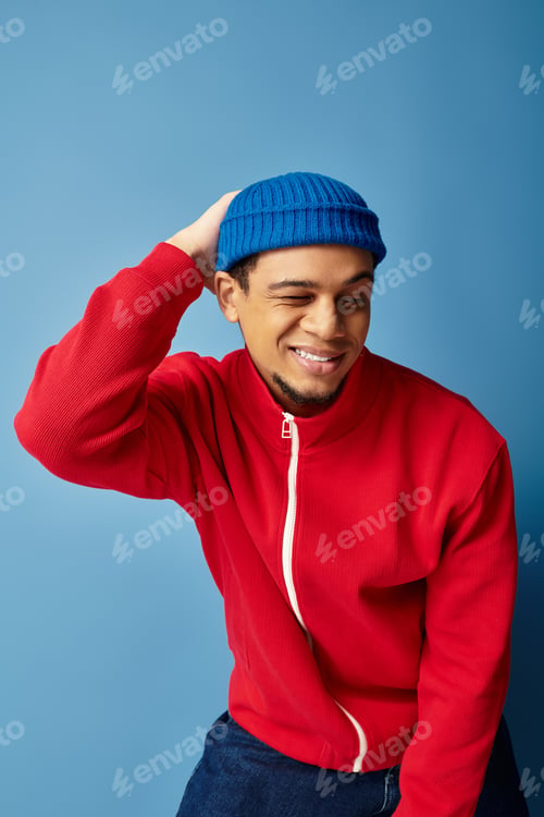 Preview: Stylish young man with a beanie hat expressing joy against a vibrant blue backdrop