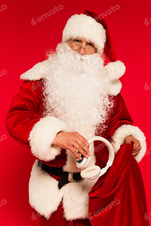 Preview: Santa claus in eyeglasses and hat putting headphones in sack isolated on red