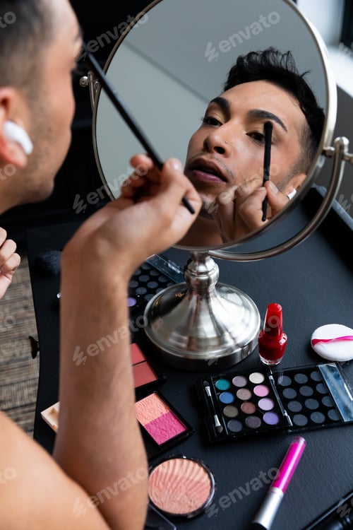 Preview: Biracial transgender man looking in mirror and putting on make-up, applying eyeshadow